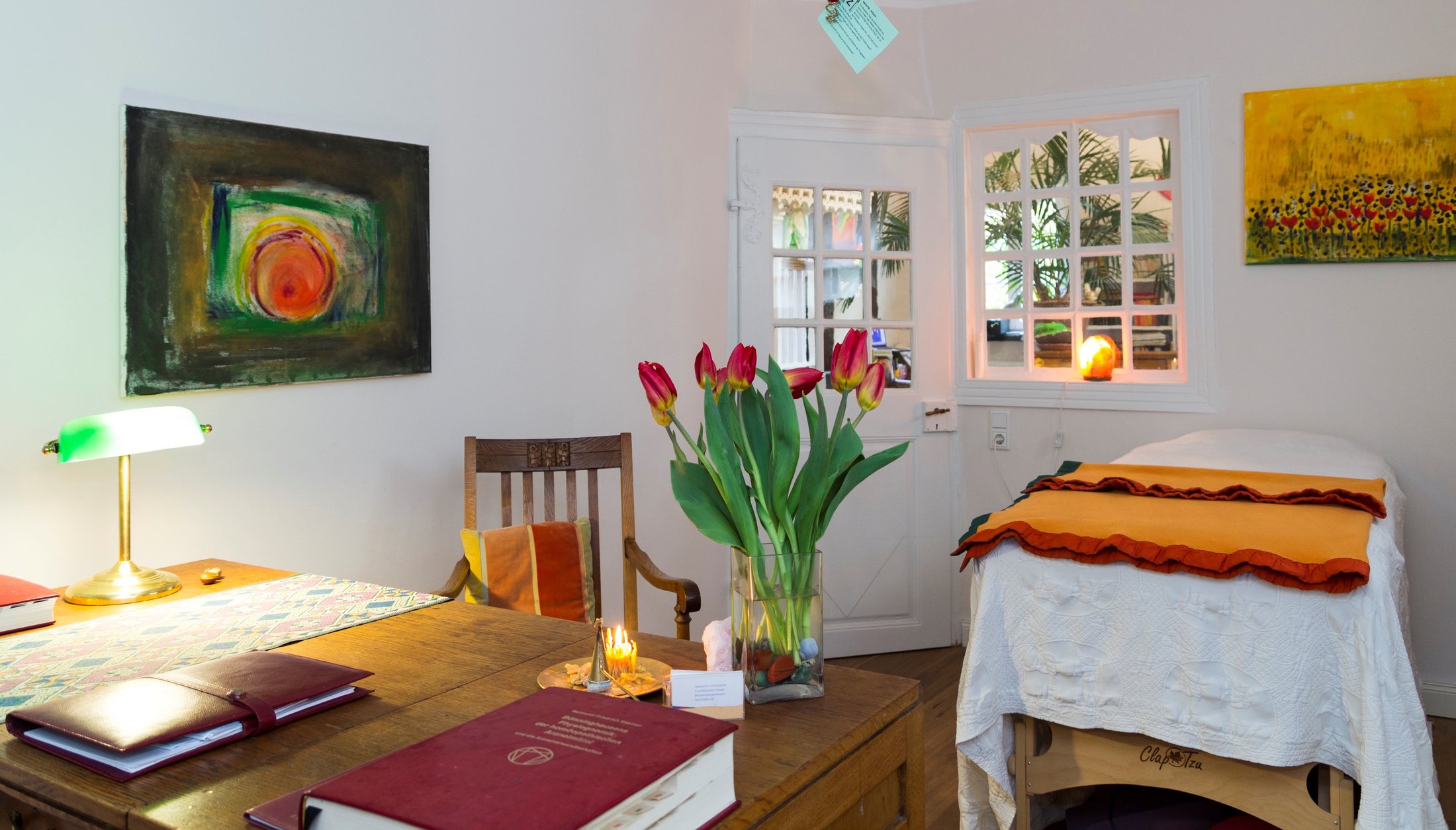 Behandlungszimmer der Praxis in Warendorf: Schreibtisch mit Tulpen und homöopathischem Nachschlagewerk, dahinter die Behandlungsliege
